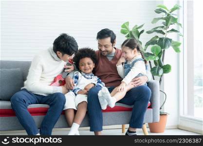 Asian young LGBTQ gay couple playing ukulele and singing with little Caucasian and African adopted kid together at home. Happy son and daughter smiling and sitting on sofa. LGBT diverse family concept