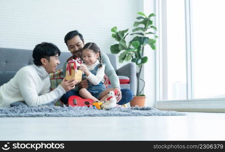 Asian young LGBTQ gay family giving gift to little adopted daughter on birthday or special day in living room at home. Caucasian kid girl opening present box with smiling and surprising face