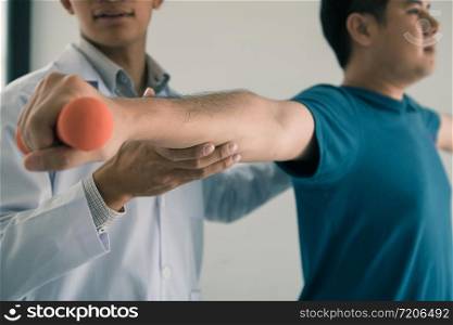 Asian young male physiotherapist helping patient with lifting dumbbells exercises in office.