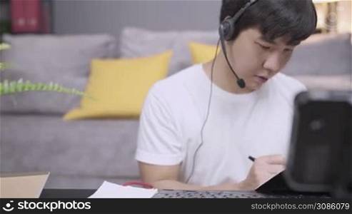Asian young mam worker wear headset call operator taking notes from client information, microphone voice call interaction with customer, live chat with staff, social distancing doing working at home