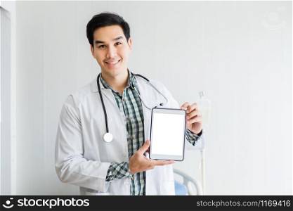 Asian Young man doctor therapeutic advising with positive emotions holding up and showing digital tablet with a blank white screen and bed in hospital background,copy space