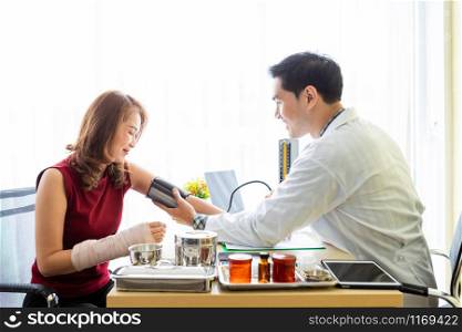Asian Young man doctor with measuring the pressure to the arm of a female patient wear arm splint with analogue pressure gauge for better healing In the room hospital background.