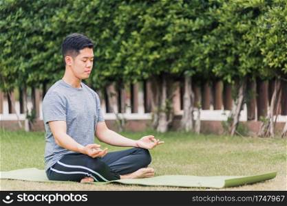 Asian young man doing yoga outdoors in meditate lotus pose sitting on green grass with closed eyes at the garden park, health care concept