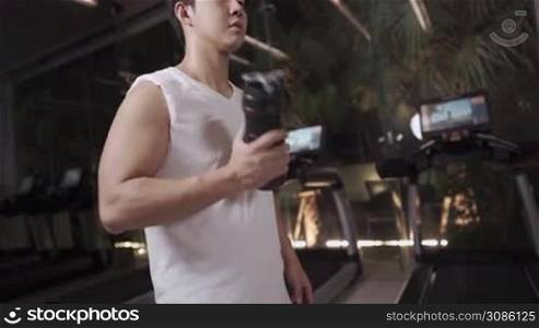 Asian young man drinking whey protein shake while resting during the sets of workout routine, rehydrate drinking water inside the gym room, body building, gaining muscle and strength, sport nutrition