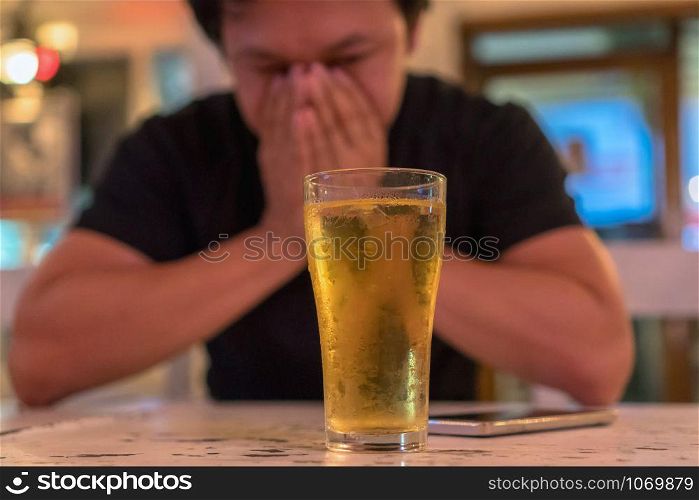 Asian young man in lonely and depressed action and holding face in hands and see glass of beer in pub and restaurant with low light place, depression and drinking concept