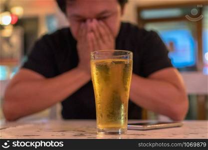 Asian young man in lonely and depressed action and holding face in hands and see glass of beer in pub and restaurant with low light place, depression and drinking concept