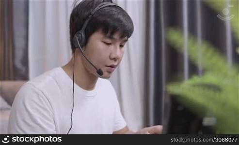 Asian young man wear headset doing work from home call operator, microphone voice call interaction with customer, live chat with staff, late night shift, casual clothing doing working at home