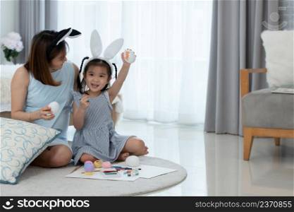 Asian young mother and little cute daughter wearing bunny ears and enjoy painting on eggs for Easter holidays while sitting on floor together at home. Happy Easter Family traditions concept