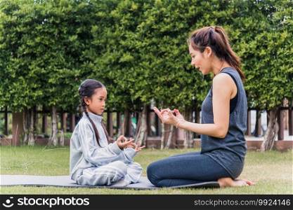 Asian young mother practicing doing yoga exercises with her daughter outdoors in meditate pose together on green grass in nature a field garden park, family sport and exercises for healthy lifestyle