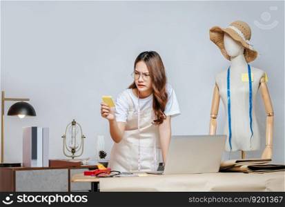 Asian young pretty female fashion designer stylish wearing eyeglasses standing and serious with paper not during working at fashion studio, full of tailoring tools and equipment