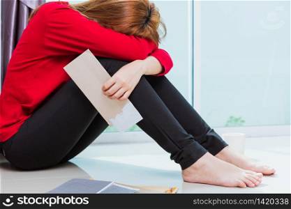Asian young sad woman hug her knee and cry wearing face mask protective unemployed quarantines disease coronavirus or COVID-19 her sitting alone on floor holding papers unpaid bills debt credit