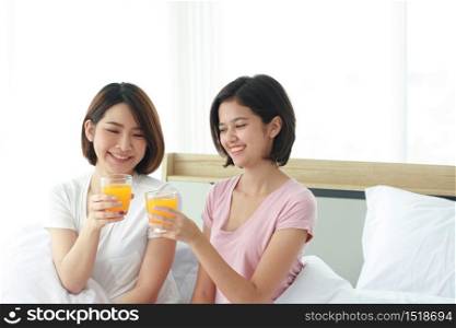 Asian young sisters lovely couple on white bed and smiling with hand holding Orange juice glass in bedroom. Holiday and healthy concept