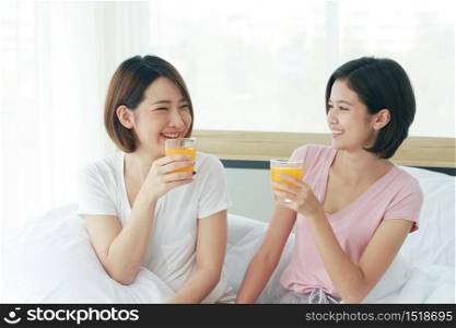 Asian young sisters lovely couple on white bed and smiling with hand holding Orange juice glass in bedroom. Holiday and healthy concept.