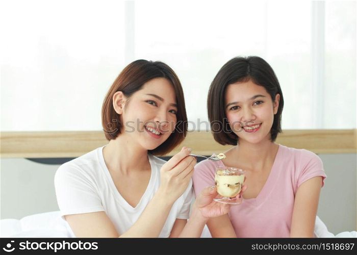 Asian young sisters lovely couple on white bed hand holding cup of cake and eating Bakery together in bedroom. Homosexual women or Lesbian in love. LGBT Asian and Holiday concept.