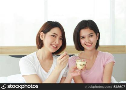 Asian young sisters lovely couple on white bed hand holding cup of cake and eating Bakery together in bedroom. Homosexual women or Lesbian in love. LGBT Asian and Holiday concept.
