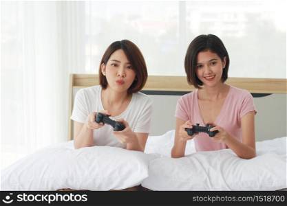 Asian young sisters lovely couple on white bed hand holding joysticks and enjoy with game play station together in bedroom. Hobby and entertainment concept