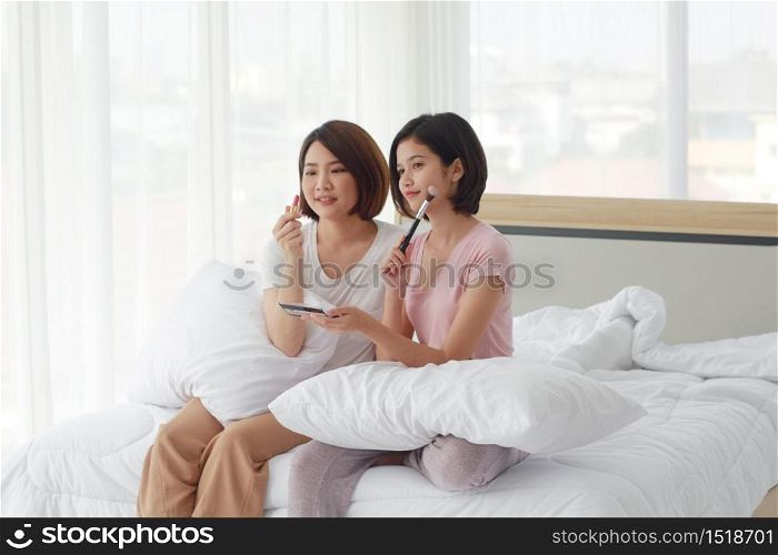Asian young sisters lovely couple on white bed smiling and make-up with lipstick together in bedroom. Homosexual women or Lesbian in love. cosmetic and holiday concept.