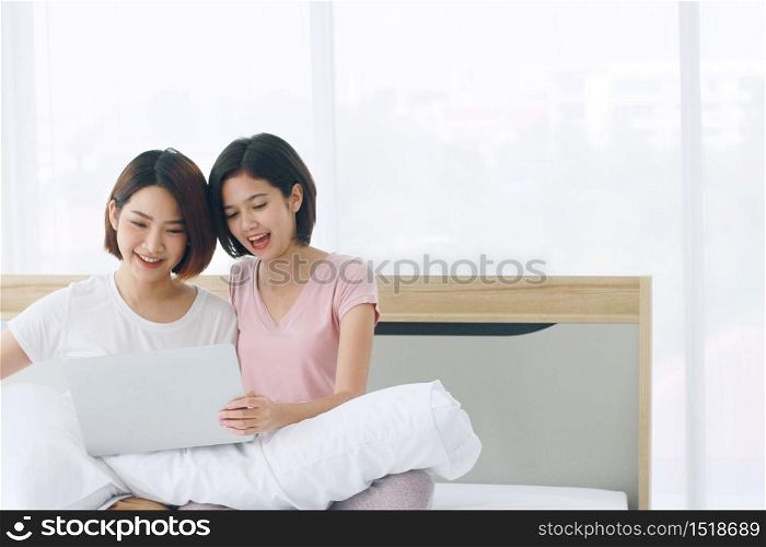 Asian young sisters lovely couple sitting on white bed and smiling together for online social with laptop in bedroom. Homosexual women or Lesbian in love. LGBT Asian and Valentine?s Day concept.