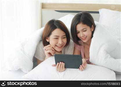 Asian young sisters lovely couple sitting on white bed and smiling together for Social online with laptop and smartphone in bedroom. Homosexual and asian Lesbian in relationship. Technology Concept
