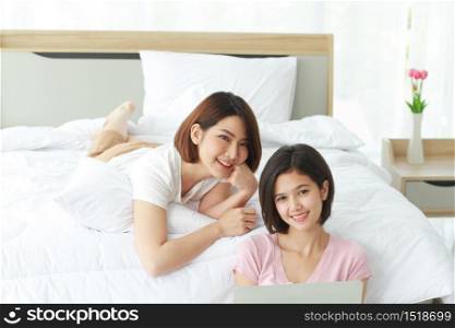 Asian young sisters lovely couple sitting on white bed and smiling together for online Social with laptop in bedroom. Homosexual women or Lesbian in love. Friendship and Holiday concept.