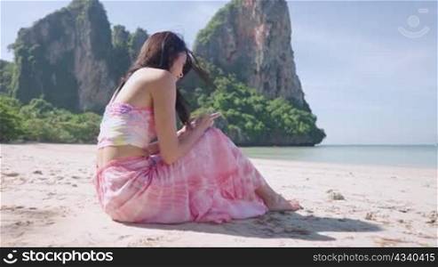 Asian young slim woman relaxing playing on phone while sitting on white beach at beautiful asia coastline, enjoying using mobile app and doing sunbath, female solo trip, remote wireless internet usage