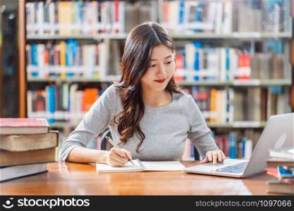Asian young Student in casual suit doing homework and using technology laptop in library of university or colleage with various book and stationary over the book shelf background, Back to school
