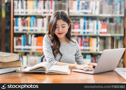 Asian young Student in casual suit doing homework and using technology laptop in library of university or colleage with various book and stationary over the book shelf background, Back to school