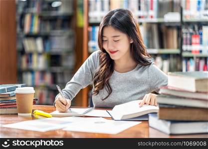 Asian young Student in casual suit reading and doing homework in library of university or colleage with various book and stationary on the wooden table over the book shelf background, Back to school