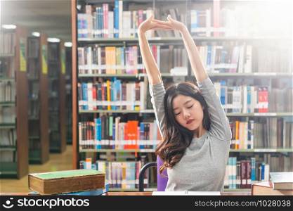 Asian young Student in casual suit reading and doing stretch oneself in library of university or colleage with various book and stationary on the wooden table over the book shelf, Back to school