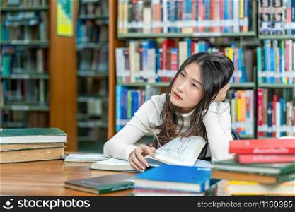 Asian young Student in casual suit reading on the wooden table with various book in library of university or colleage over the book shelf, Back to school concept