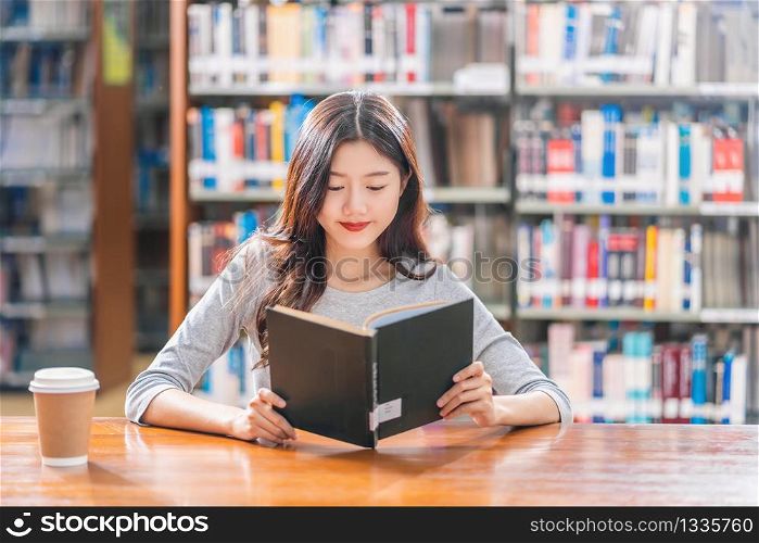 Asian young Student in casual suit reading the book with a cup of coffee in library of university or colleage on the wooden table over the book shelf background, Back to school concept