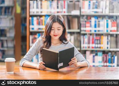 Asian young Student in casual suit reading the book with a cup of coffee in library of university or colleage on the wooden table over the book shelf background, Back to school concept