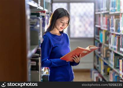 Asian young Student in casual suit standing and reading the book at book shelf in library of university or colleage with various book background, Back to school concept