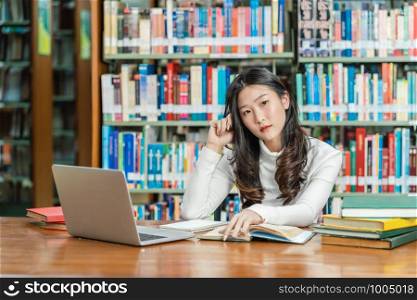 Asian young Student in casual suit thinking when doing homework and using technology laptop in library of university or colleage with various book and stationary over the book shelf, Back to school