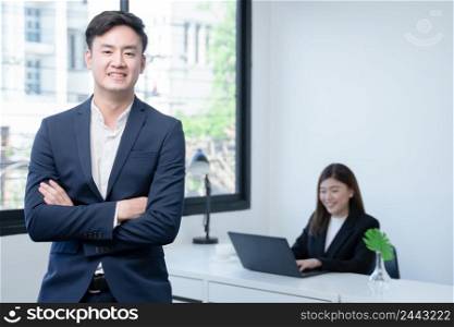 Asian young successful businessman wear formal suit smiling with confident and standing with crossed arm at office with his colleagues woman working on laptop on blur background. White background