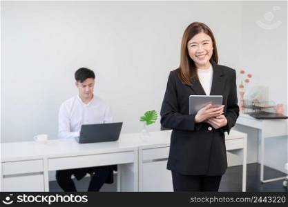 Asian young successful businesswoman wear formal suit standing and holding tablet smiling with confident at office with her colleagues man working on laptop on blur background. White background
