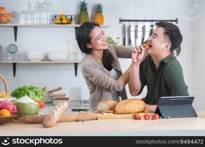 Asian young sweet couple cooking breakfast together in home kitchen. Beautiful wife smiling embracing her husband from back while he eating strawberry and working on tablet. Family lifestyle
