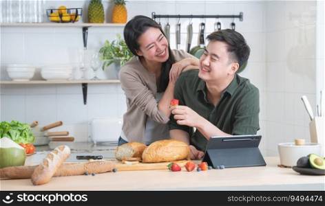 Asian young sweet couple cooking breakfast together in home kitchen. Beautiful wife smiling embracing her husband from back while he eating strawberry and working on tablet. Family lifestyle