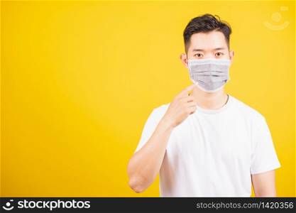 Asian young teen man wearing surgical hygienic protective face mask against coronavirus and pointing finger to mask on face, studio shot isolated white background, COVID-19 medical concept