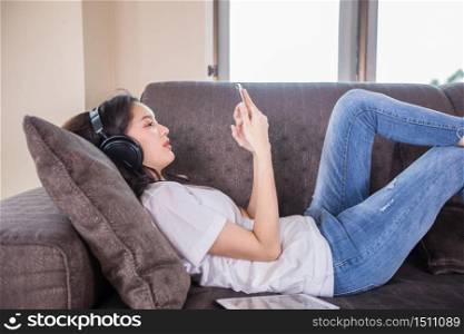 Asian young woman happy cheerful cute beautiful wears a headset for listening to music from a mobile phone and she is playing smartphone and lying on the sofa at home,