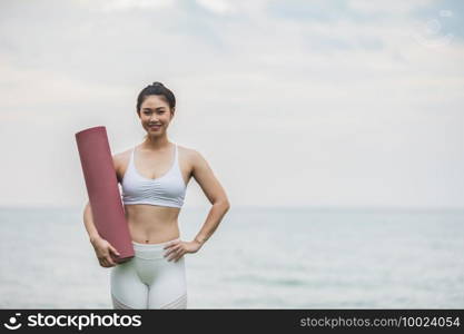 Asian young woman holding yoga mat and smile on the beach,ready to practice yoga with happiness feeling