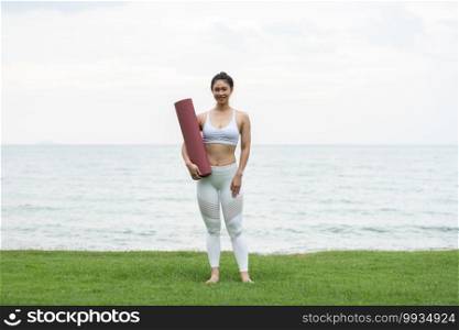 Asian young woman holding yoga mat and smile on the beach,ready to practice yoga with happiness feeling