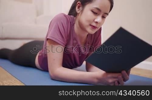 Asian young woman lying down on yoga mat reading book at home lockdown period, quarantine covid-19, after home work out exercise, relaxing hour on leisure free time, well being calm relaxation