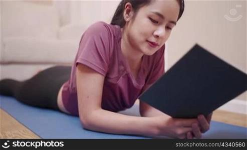 Asian young woman lying down on yoga mat reading book at home lockdown period, quarantine covid-19, after home work out exercise, relaxing hour on leisure free time, well being calm relaxation