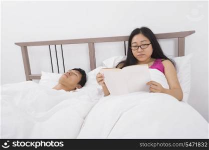 Asian young woman reading book by man sleeping in bed
