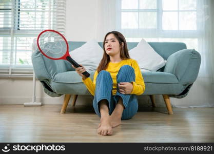 Asian young woman sitting floor using mosquito swatter or electric net racket, female killing mosquitoes she holding fly swatter like weapon in living room at home, Insect killer