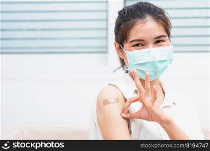 Asian young woman smile she’s have adhesive plaster on arm her vaccinated and showing OK finger sign after getting immunity vaccine COVID-19 prevent