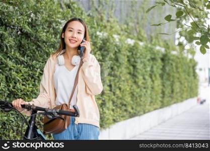 Asian young woman walking in countryside outdoor with her bicycle she using mobile phone talking with friend, Happy female stand on city street with bike using smartphone for summer sightseeing walk