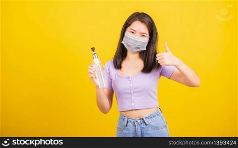 Asian young woman wearing face mask protective against coronavirus or COVID-19 virus or filter dust, air pollution her hold alcohol gel show finger thumb up for good sign, isolated yellow background