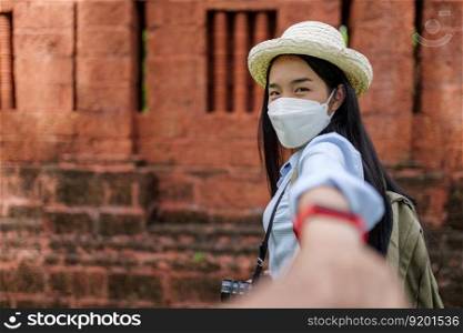 Asian young woman wearing protection mask and holding hand of crop person while walking, blurred background with ancient site, New normal of traveller